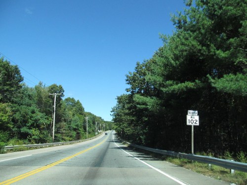 A clear road stretches ahead with a sign indicating "North RI 102," surrounded by trees under a blue sky.