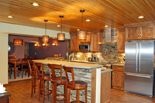 Modern kitchen with wooden cabinets, stone accents, pendant lights, and a bar with stools, featuring an open dining area.