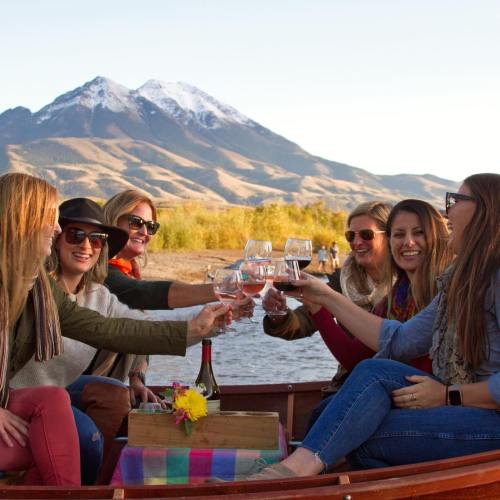 A group of six women cheers with wine glasses on a boat, with mountains and a clear sky in the background.