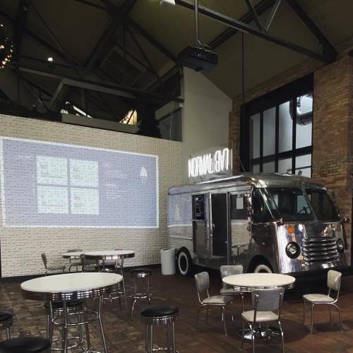 A silver food truck inside a spacious venue with tables, a projector screen, and a modern industrial design.