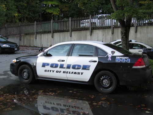 A Belfast police car parked beside a puddle, featuring a black and white design with "City of Belfast" and emergency number.