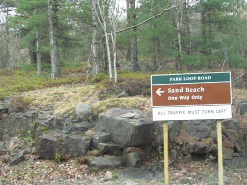 A brown and white sign indicating "Sand Beach" with a left arrow, surrounded by trees and rocky terrain.