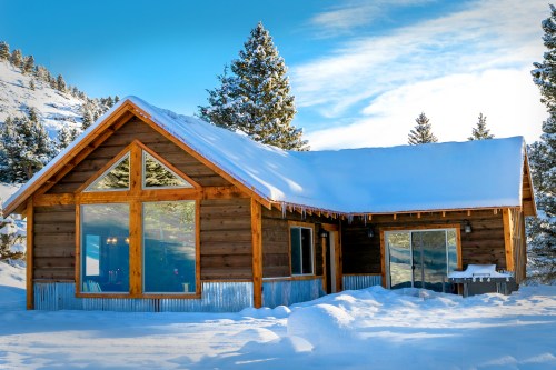 A cozy wooden cabin covered in snow, surrounded by evergreen trees under a bright blue sky.