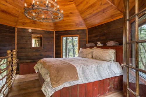 Cozy bedroom in a wooden cabin with a chandelier, large windows, and a rustic bedspread.