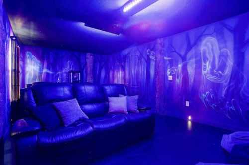 A dimly lit room with blue walls featuring ghostly forest murals and a black leather couch with pillows.