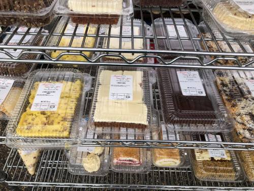 A display of assorted cakes and desserts in clear plastic containers on a metal rack.