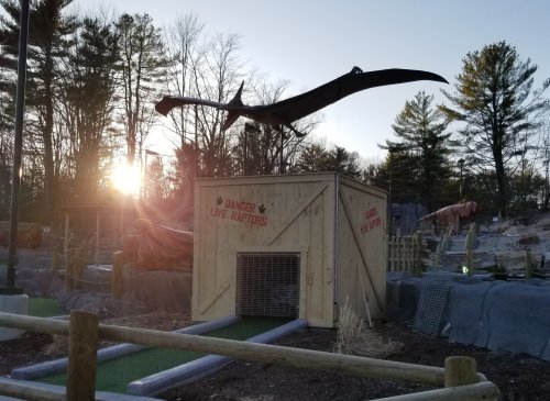 A wooden enclosure labeled "Danger: Live Raptors" with a dinosaur silhouette above, set in a wooded area at sunset.