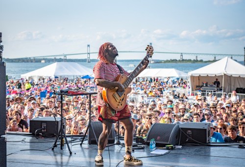 A musician performs on stage with a guitar, facing a large crowd and a scenic waterfront backdrop.
