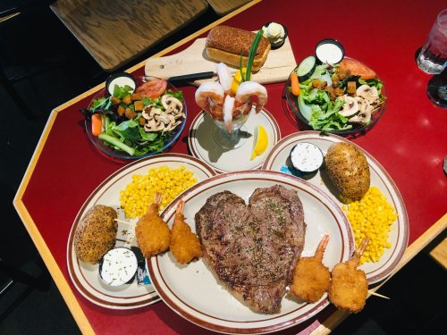 A hearty meal featuring a large steak, shrimp, salads, baked potatoes, corn, and a seafood cocktail on a red table.