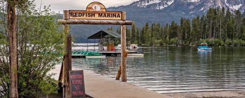 Sign for Redfish Marina at a serene lake, with boats and mountains in the background.