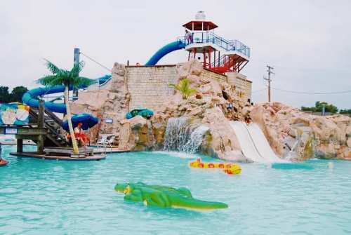 A water park scene featuring a large slide, a rock formation, and a green alligator float in a pool.