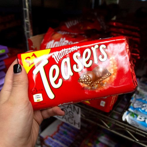 A hand holding a red package of Maltesers Teasers chocolate in a store aisle. Price tag shows $1.