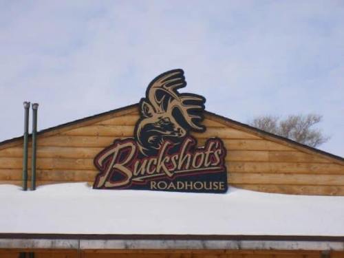 Sign for Buckshots Roadhouse on a wooden building, with a moose graphic above the name, set against a snowy background.