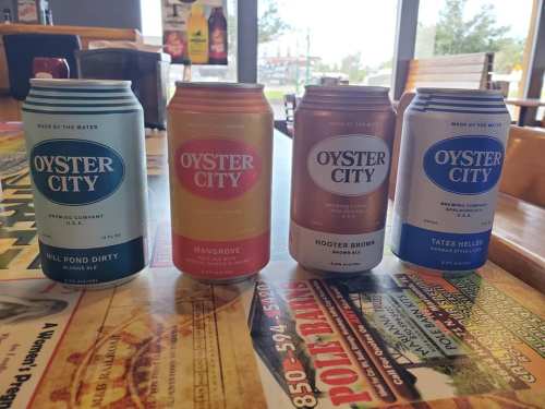 Four cans of Oyster City beer in various flavors displayed on a table, with a restaurant background.