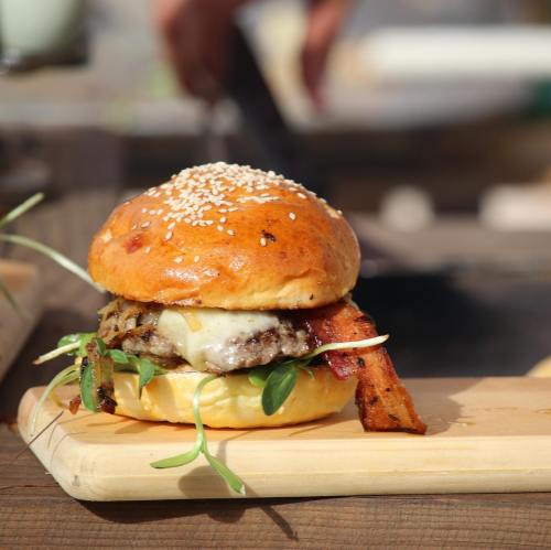 A delicious burger with melted cheese, bacon, and fresh greens on a sesame seed bun, served on a wooden board.