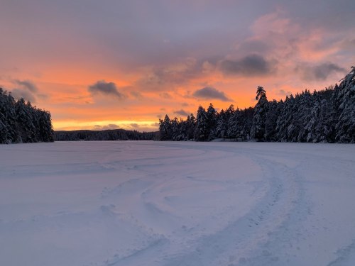 A snowy landscape at sunset, with vibrant orange and purple skies and snow-covered trees lining the horizon.