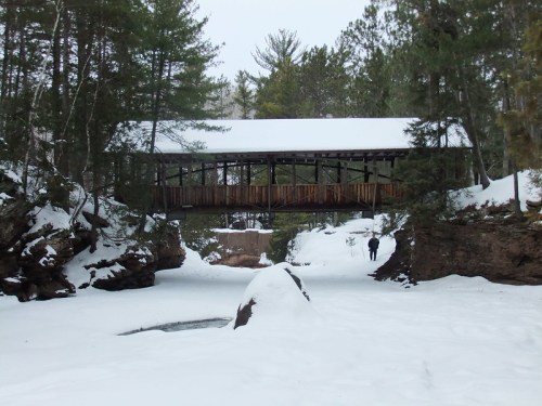 A snow-covered landscape featuring a wooden bridge over a frozen stream, surrounded by trees and rocky terrain.