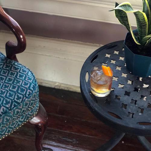 A glass of cocktail with an orange slice sits on a decorative metal table next to a potted plant and a patterned chair.