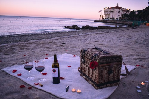 A romantic beach picnic setup with wine glasses, a bottle, a picnic basket, rose petals, and candles at sunset.