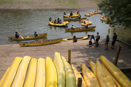 A group of people in yellow kayaks on a river, with others preparing on a wooden dock nearby.