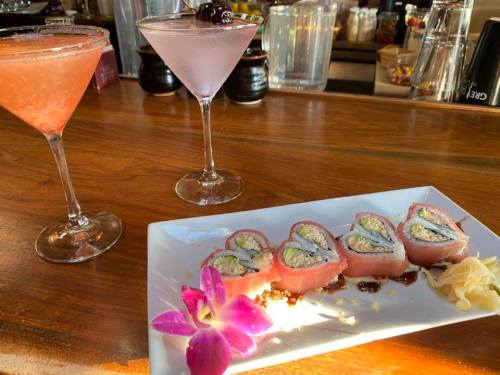 A plate of sushi rolls with a pink flower, accompanied by two colorful cocktails on a wooden bar.