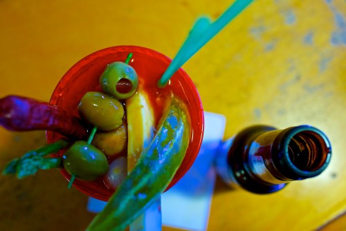 A vibrant cocktail garnished with olives, pickles, and a chili pepper, next to a beer bottle on a wooden table.
