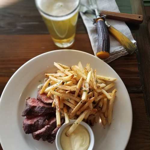 A plate of sliced steak with crispy fries and dipping sauce, accompanied by a glass of beer and a knife on the side.