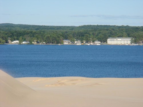 A sandy dune in the foreground with a view of a calm lake and a tree-lined shore in the background.