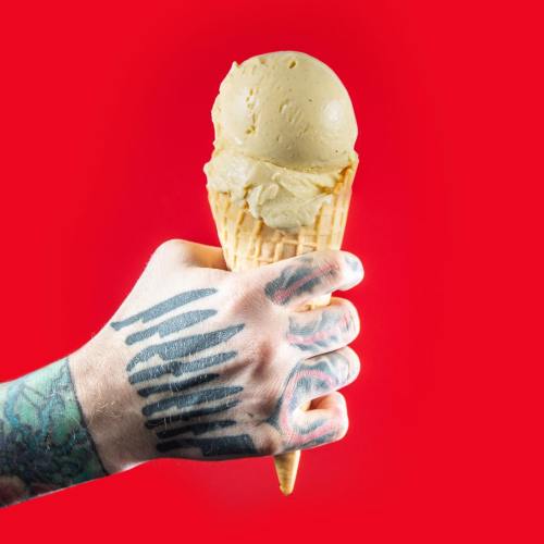 A hand with tattoos holds an ice cream cone against a bright red background.