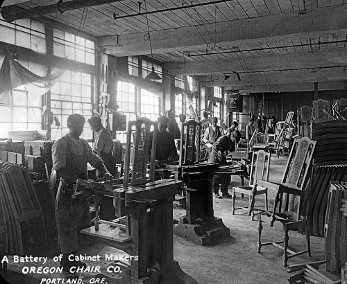 Black and white photo of workers in a furniture factory, crafting chairs with tools and wooden frames around them.