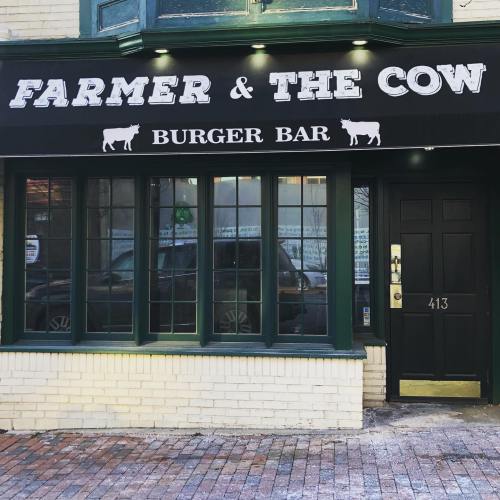 Exterior of "Farmer & The Cow Burger Bar," featuring a black sign with white lettering and large windows.