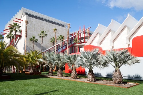 Modern architecture featuring palm trees, red accents, and a green lawn under a clear blue sky.