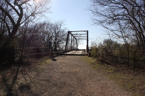 A gravel path leads to an old metal bridge surrounded by trees under a clear blue sky.