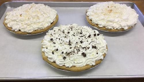 Three cream pies on a baking tray, topped with whipped cream and chocolate shavings, with one pie lightly toasted.