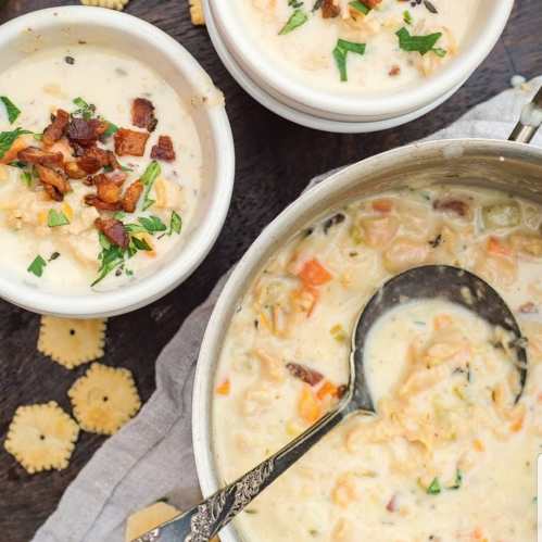 Creamy soup in a pot with two bowls, topped with bacon and herbs, surrounded by crackers on a wooden surface.