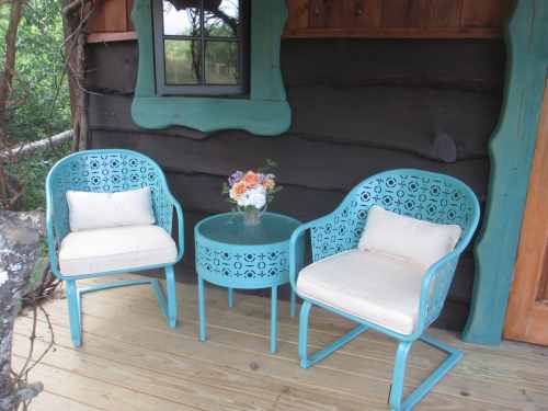 Two turquoise chairs with cushions and a small table, adorned with a vase of flowers, set against a wooden wall.