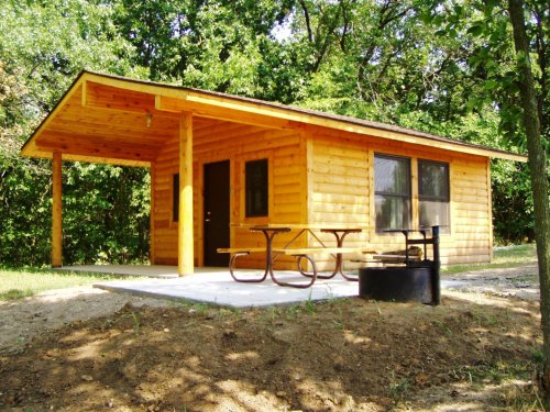 A wooden cabin with a porch, picnic table, and fire pit, surrounded by trees in a natural setting.