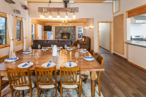 A spacious dining area with a large wooden table set for dinner, surrounded by windows and a cozy living space.