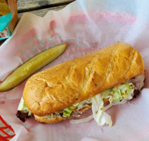A sub sandwich with lettuce and meat, served on a red and white checkered paper, alongside a pickle.