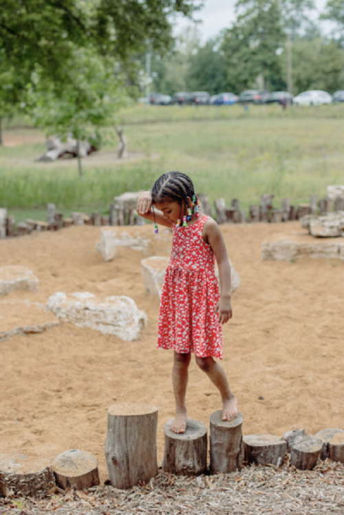 A young girl in a red dress balances on wooden stumps in a sandy playground, surrounded by greenery.