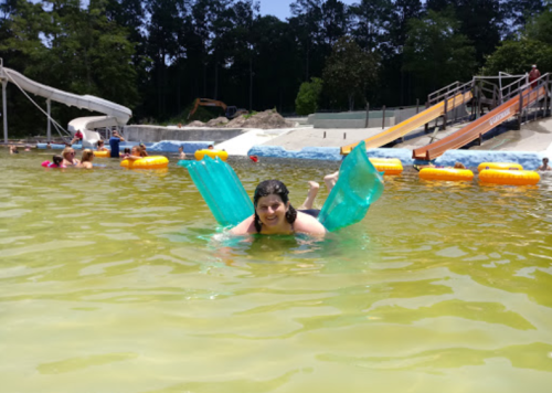 A person swims in a pool with green floaties, surrounded by other people and water slides in the background.