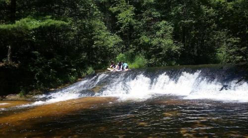Three people sit on a rocky ledge by a waterfall, surrounded by lush green trees and flowing water.