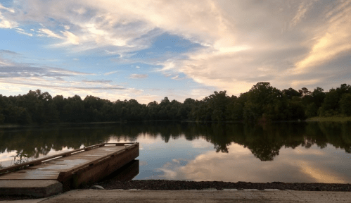 A serene lake at sunset, with a wooden dock and trees reflecting in the calm water under a colorful sky.