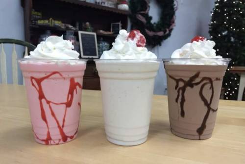 Three milkshakes in clear cups: pink with red drizzle, white, and brown with chocolate drizzle, all topped with whipped cream and cherries.