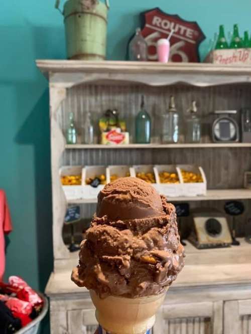 A close-up of a chocolate ice cream cone in front of a rustic shelf with vintage decor and snacks.