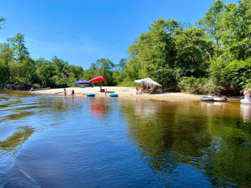 A serene river scene with people enjoying a sandy beach, tents, and inflatable tubes under a clear blue sky.