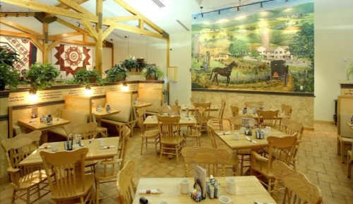 Cozy restaurant interior with wooden tables, chairs, and a mural of a countryside scene on the wall.
