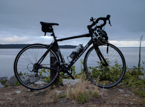 A black road bike parked by a calm body of water, surrounded by grass and rocks under a cloudy sky.