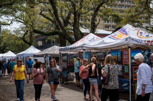 A bustling outdoor art market with people browsing colorful tents and artwork under trees.