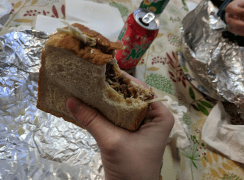 A hand holding a sandwich filled with meat and toppings, with a can of soda and wrapped food in the background.